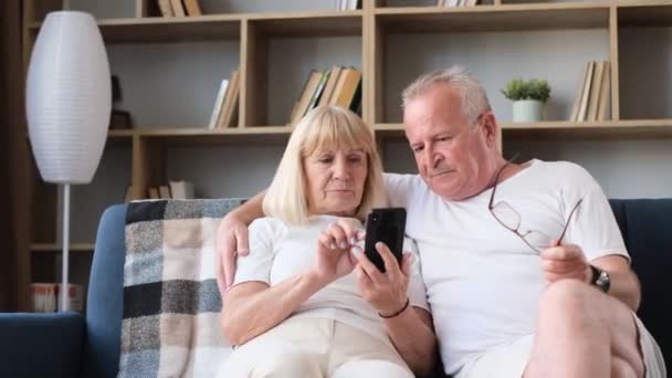 Modern retired couple sitting on sofa and using mobile apps. Grandfather and grandmother with a smartphone in their hands. Modern mobile phones - 映像、動画