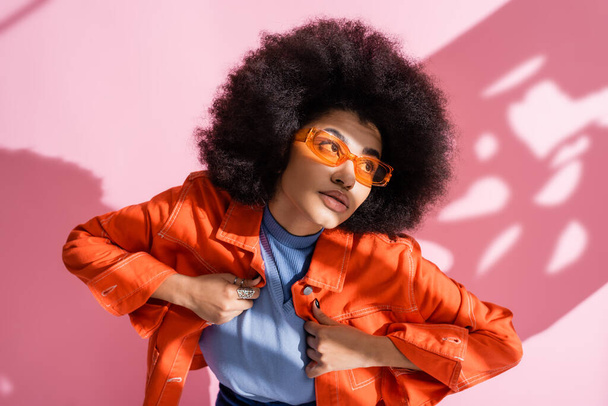 göndör afro-amerikai nő stílusos narancssárga napszemüveg és divatos ruhát pózol rózsaszín  - Fotó, kép