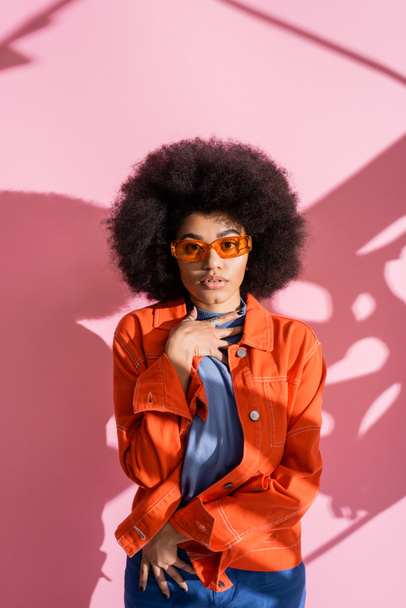curly african american woman in stylish orange sunglasses and trendy outfit looking at camera on pink  - Zdjęcie, obraz