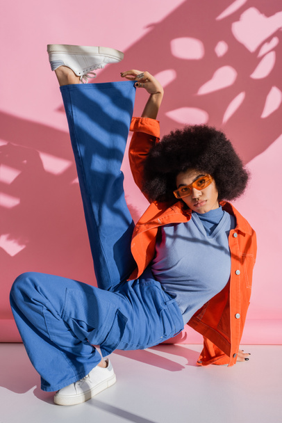 comprimento total de mulher americana africana encaracolado em óculos de sol laranja elegantes e roupa na moda posando em rosa  - Foto, Imagem