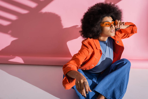 curly african american woman in trendy outfit adjusting orange sunglasses on pink  - Foto, afbeelding