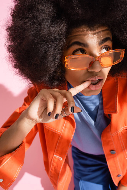 σγουρή Αφροαμερικανή με πορτοκαλί γυαλιά ηλίου που δαγκώνει το δάχτυλο και κοιτάει αλλού το ροζ.  - Φωτογραφία, εικόνα