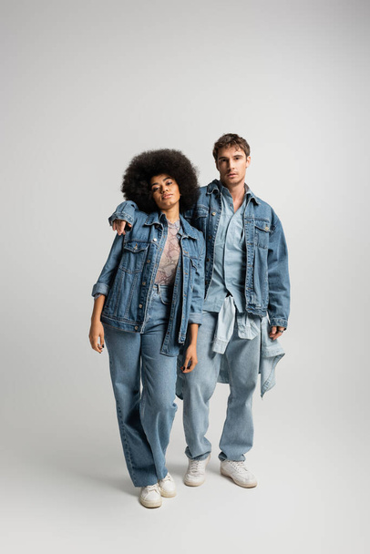 full length of young man in denim outfit hugging african american woman on grey  - Foto, imagen