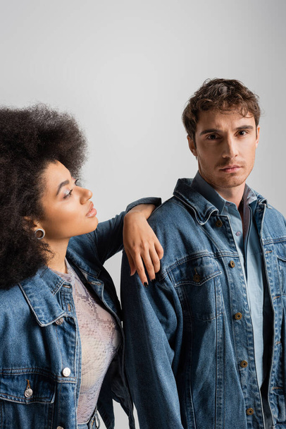 curly african american woman leaning on shoulder of young man in denim outfit isolated on grey  - Φωτογραφία, εικόνα