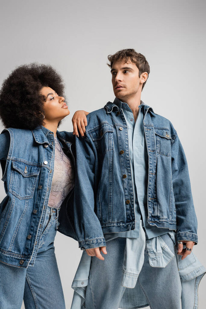 lockiges afrikanisch-amerikanisches Model stützt sich auf die Schulter eines jungen Mannes im Jeans-Outfit isoliert auf grau  - Foto, Bild