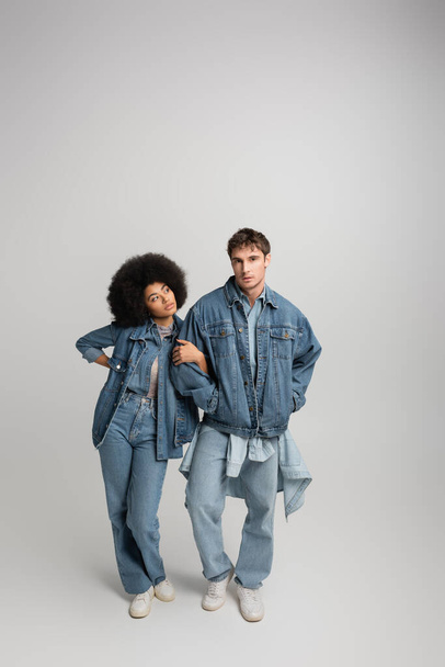 full length of young man in denim outfit posing with hands in pockets near curly african american woman on grey  - Valokuva, kuva