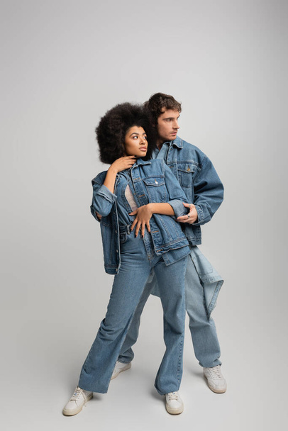 full length of young multiethnic couple in denim outfit hugging and looking away on grey  - 写真・画像