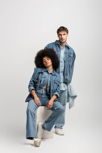 full length of curly african american woman sitting near man in denim outfit on grey  - Foto, Bild