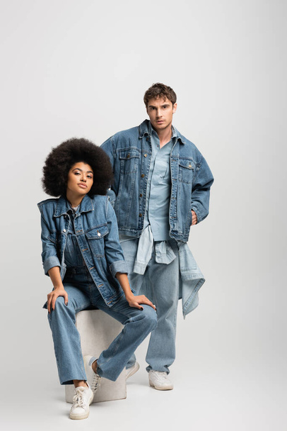 full length of curly african american woman sitting near man in denim outfit posing with hands in pockets on grey  - Zdjęcie, obraz