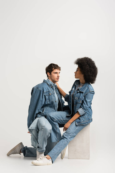 full length of african american woman sitting and looking at man in denim outfit on grey  - 写真・画像