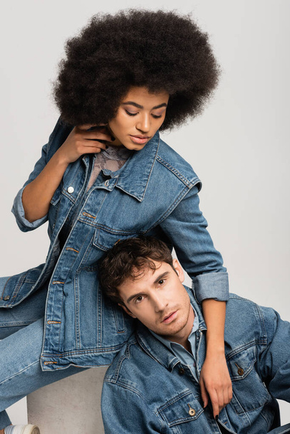 brunette african american woman sitting and looking at man in denim outfit isolated on grey  - Foto, imagen