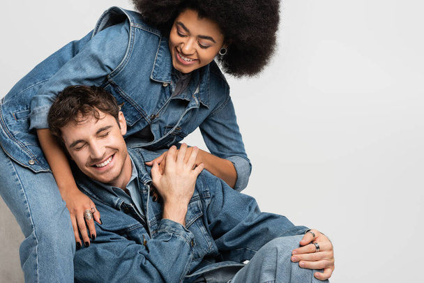 щаслива афроамериканка сидить і дивиться на веселого чоловіка в джинсовому вбранні ізольовано на сірому
  - Фото, зображення