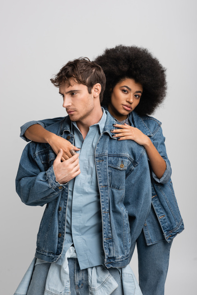 ricci modello africano americano guardando la fotocamera mentre abbraccia l'uomo elegante in abiti di jeans isolati su grigio  - Foto, immagini