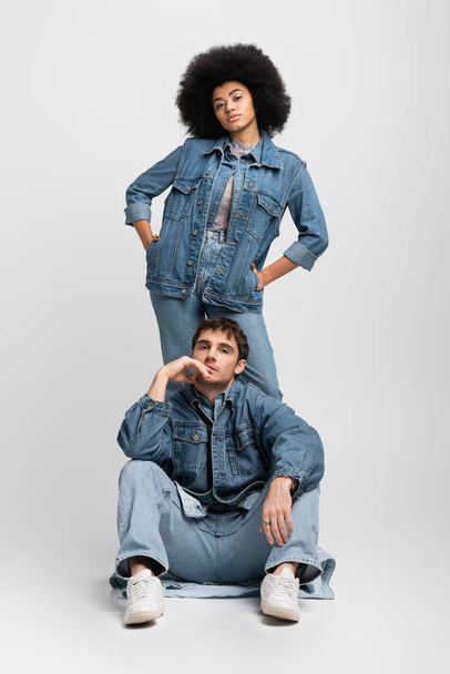 full length of curly african american woman standing with hands in pockets behind man in denim outfit on grey - 写真・画像
