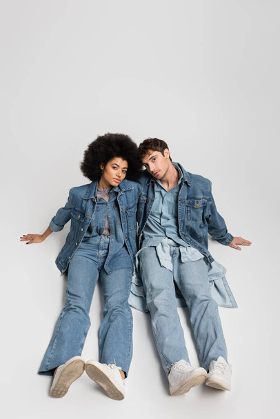 full length of stylish interracial couple in denim clothes sitting in sneakers on grey  - Foto, imagen