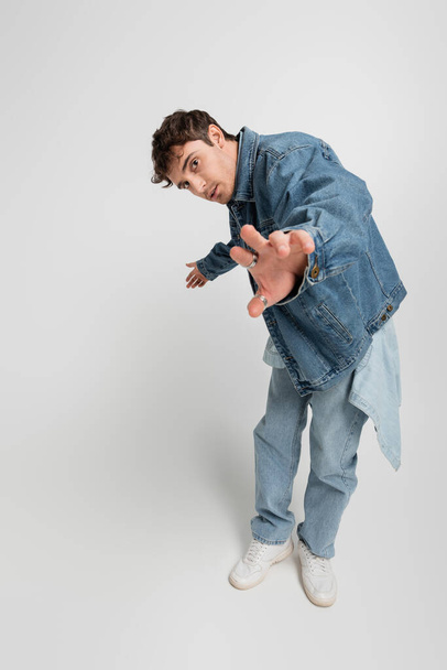 полная длина стильного молодого человека в джинсовой куртке и белых кроссовках, стоящих с протянутой рукой на сером  - Фото, изображение