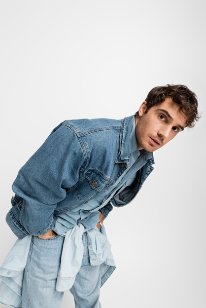 stylish young man in denim clothes posing with hands in pockets and looking at camera isolated on grey  - Фото, изображение