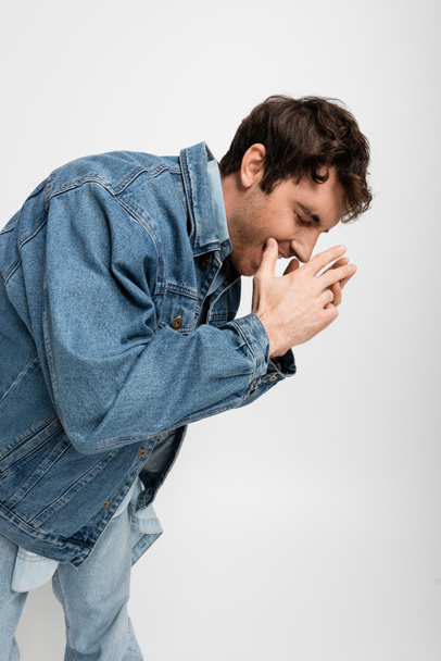 stylový mladý muž v džínové bundě se zasmátýma zavřenýma očima izolovaný na šedi  - Fotografie, Obrázek