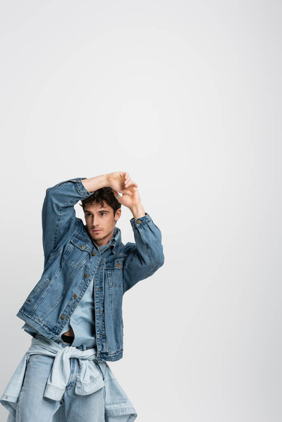 стильный молодой человек в джинсовой куртке и джинсах позирует с руками над головой изолированы на сером  - Фото, изображение