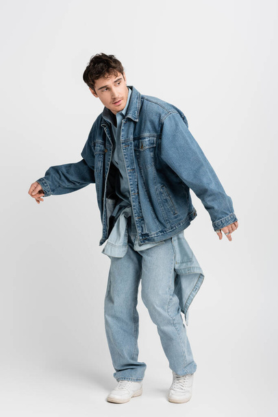 full length of stylish young man in denim jacket and jeans posing while looking away on grey  - 写真・画像