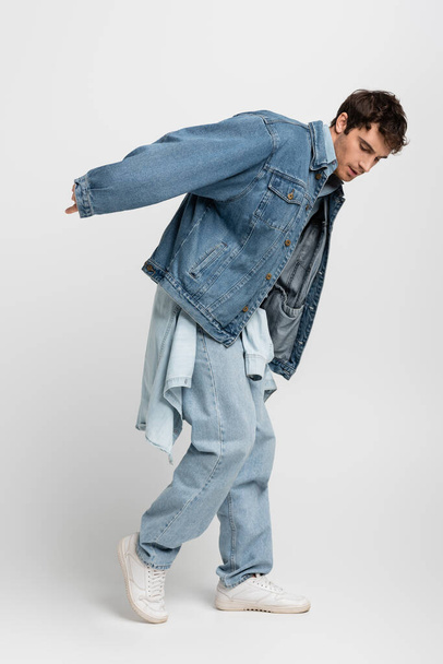 полная длина стильный человек в джинсовой куртке и джинсах позируя, глядя вниз на серый  - Фото, изображение