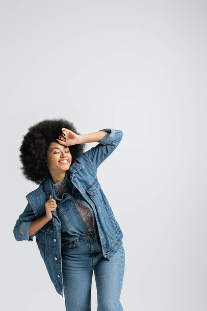 pleased african american woman in denim jacket smiling while closed eyes isolated on grey  - Foto, immagini