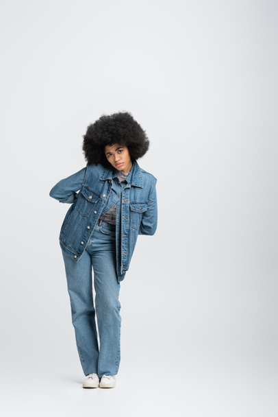 full length of curly african american woman in denim outfit looking at camera while standing on grey  - Valokuva, kuva