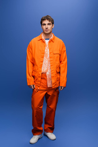 Full length of stylish man in orange jacket and overall standing on blue background - Foto, Bild