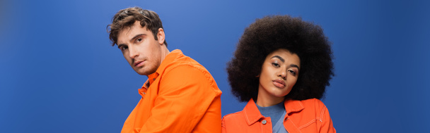 Multiethnic couple in orange jackets looking at camera isolated on blue, banner  - Foto, Imagem