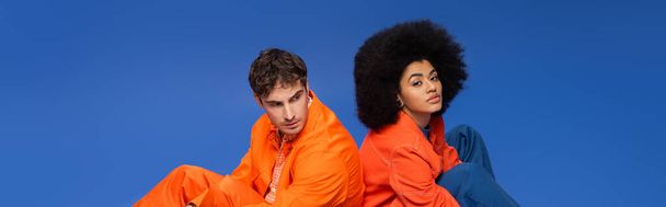 Modèle afro-américain regardant la caméra près de l'homme en vêtements orange isolé sur bleu, bannière  - Photo, image