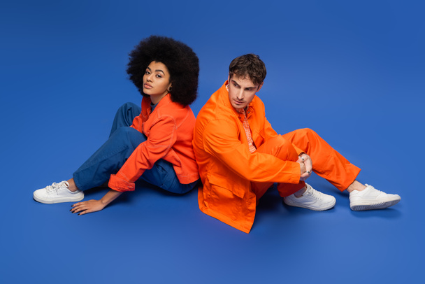Stylish multiethnic couple in jackets sitting on blue background - Foto, Imagen
