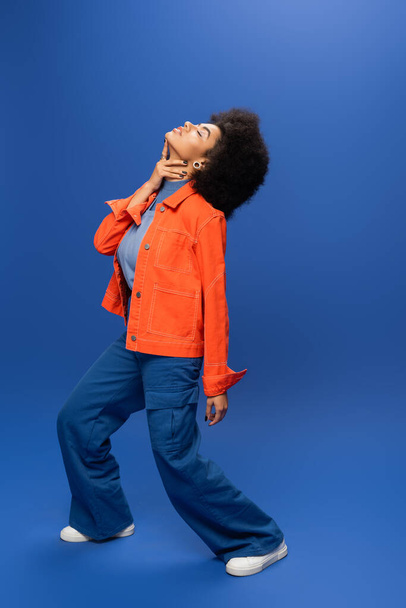 Fashionable african american woman in jacket and pants touching neck on blue background - Photo, Image
