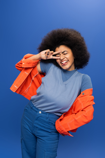 Cheerful african american woman in bright outfit showing peace sign on blue background - Valokuva, kuva
