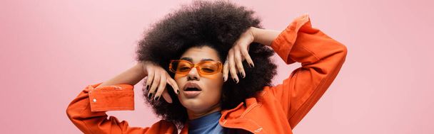Stylish african american model in orange jacket and sunglasses isolated on pink, banner  - Foto, immagini