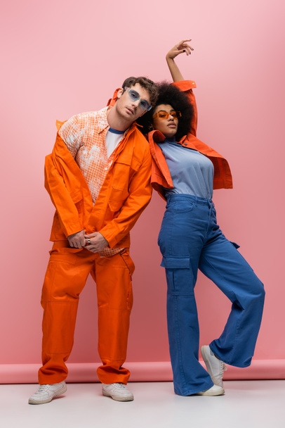 Trendy interracial couple in sunglasses posing and looking at camera on pink background  - Foto, imagen