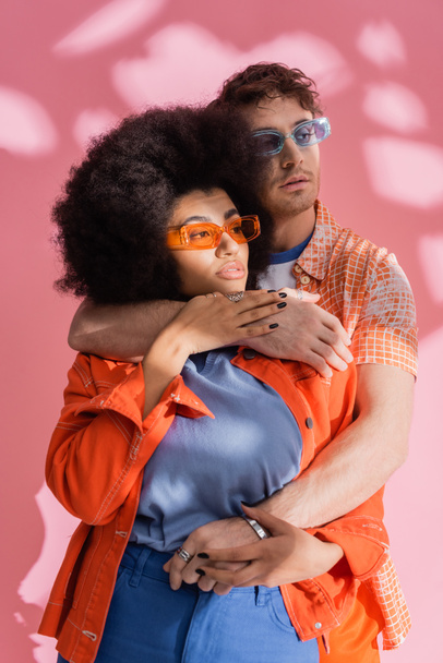 Stylish brunette man hugging african american woman on pink background with shadow  - Fotografie, Obrázek