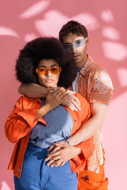 Brunette man hugging stylish african american woman in sunglasses on pink background with shadow  - Foto, Imagen