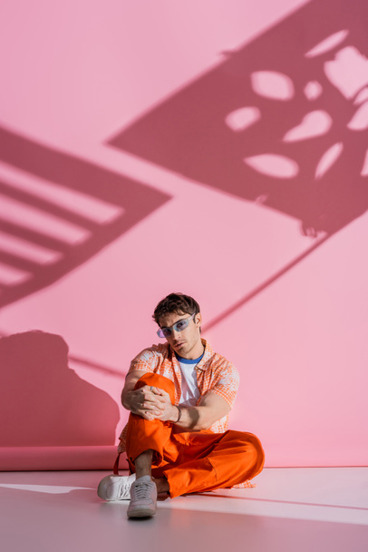 Trendy giovane uomo in occhiali da sole seduto su sfondo rosa con ombra  - Foto, immagini