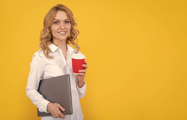 positieve blonde vrouw met koffiekop en laptop op gele achtergrond. - Foto, afbeelding