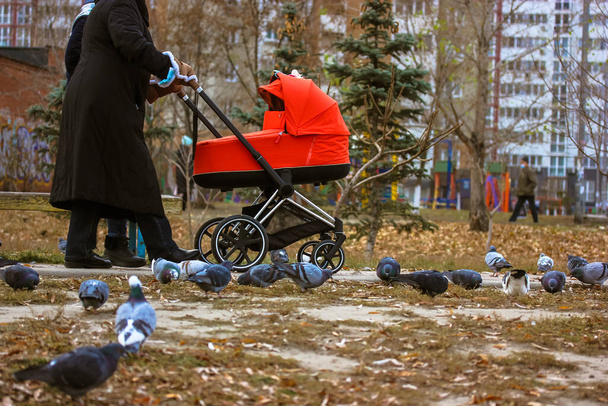 Kobieta bez twarzy z czerwonym wózkiem spaceru z niemowlęciem na ulicy miasta w zimny jesień lub zimowy dzień. Macierzyństwo, opieka nad dziećmi, koncepcja rodzicielstwa. Matka na świeżym powietrzu z małym nowonarodzonym maluchem.  - Zdjęcie, obraz