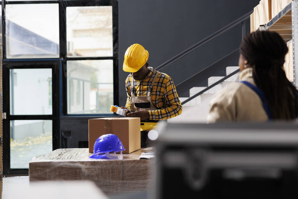 Αφροαμερικάνος που πακετάρει πακέτα πελατών σε ταχυδρομική αποθήκη. Υπάλληλος βιομηχανικής αποθήκης φορώντας σκληρό καπέλο σφράγιση κουτί από χαρτόνι με τη χρήση κολλητική ταινία διανομέα - Φωτογραφία, εικόνα
