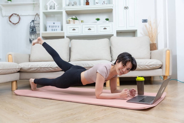 Вписується азіатська жінка робить йогу дошку і дивиться онлайн підручники з ноутбука. навчання у вітальні
 - Фото, зображення