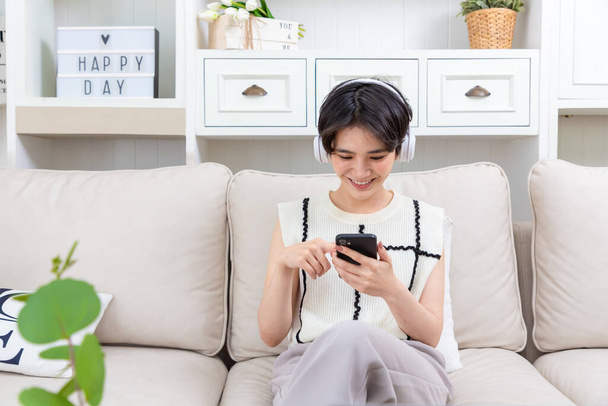 szczęśliwy Azji dziewczyna noszenie słuchawki oglądanie i słuchanie filmy na laptop siedzi na kanapie w salonie. - Zdjęcie, obraz