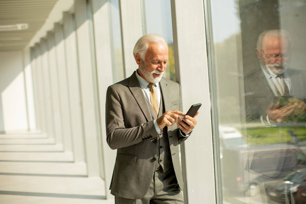 Starszy biznesmen stoi w biurze, skupiony na telefonie komórkowym. Jest ubrany w strój formalny, emanuje pewnością siebie i profesjonalizmem - Zdjęcie, obraz