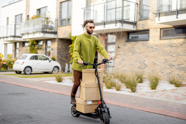 Mann fährt Elektroroller, liefert Kartons auf Straße in Wohngebiet aus Konzept der Nachhaltigkeit, Lieferung und umweltfreundlicher moderner Lebensstil - Foto, Bild