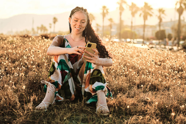 Woman with vitiligo using her mobile phone while sitting outdoors on grass in park. Technology concept. - Foto, imagen