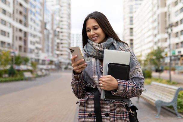 Une femme d'affaires en route pour travailler avec un ordinateur portable dans ses mains regarde l'écran du téléphone avec un sourire. - Photo, image