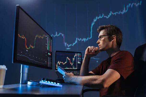 Side view of concentrated crypto trader sitting in front of computers, making professional analysis of candlestick chart, creating strategy, holding pen and notepad, looking at monitor, touching chin - Foto, afbeelding