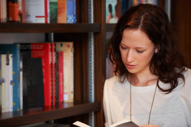 Βυθισμένος σε ένα νέο μυθιστόρημα. Μια νεαρή γυναίκα διαβάζει ένα βιβλίο ενώ ακουμπάει σε κάποια ράφια. - Φωτογραφία, εικόνα