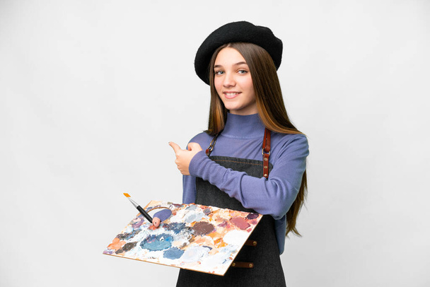 Молодой художник держит палитру на изолированном белом фоне, указывая назад - Фото, изображение